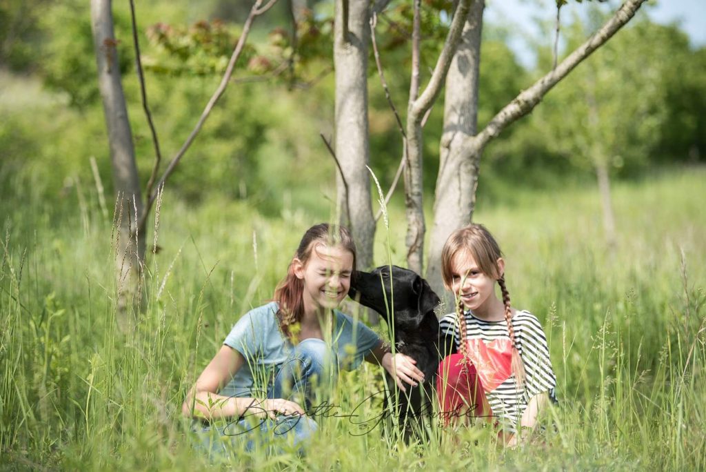 Kinder Hundezone. Zwei junge Mädchen hockeln in der Wiese mit ihrem schwarzen Hund, der einem Mädchen ein Küsschen gibt.